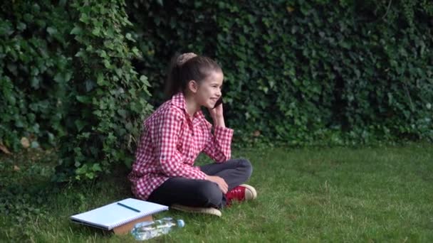 Une jeune femme caucasienne de la génération numérique est assise dans le parc sur l'herbe verte, tient un smartphone et communique au téléphone. — Video