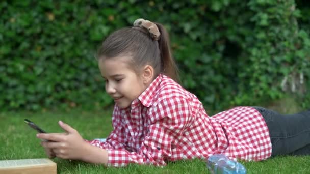 Kleine mooie blanke dame ligt op het groene gras in het park en communiceren met vrienden via de applicatie op de smartphone. — Stockvideo