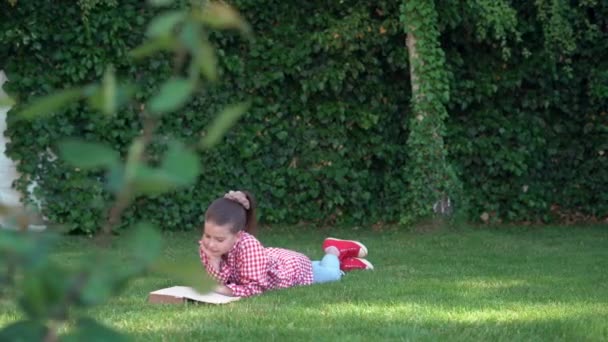 En vacker leende skolflicka i avslappnade kläder, en röd skjorta, liggande på gräset och läsa en bok, mot bakgrund av en grön sommarpark. — Stockvideo