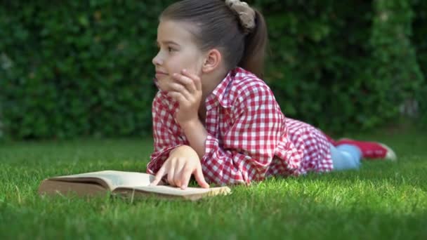 Een jong blank schoolmeisje ligt op het groene gras en leest een boek. Kijk om je heen en droom. Kopieerruimte. — Stockvideo