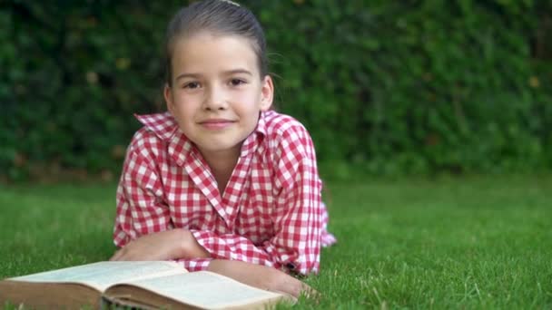 A young Caucasian schoolgirl girl lies on the green grass and reads a book. looks into the camera with a confidential smile. Copy space. — Stock Video