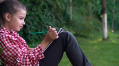 Büyük liseli bir kızın portresi parktaki yeşil çimlerin üzerinde bir ağacın yanında oturuyor ve kalemle kağıda kelimeleri yazıyor..