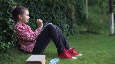 Parkta açık havada düşünmek. Kırmızı gömlekli genç beyaz bir kız parktaki yeşil çimlerin üzerinde bir ağacın yanında oturmuş bir şeyler düşünüyor ve düşüncelerini kalemle bir deftere yazıyor..