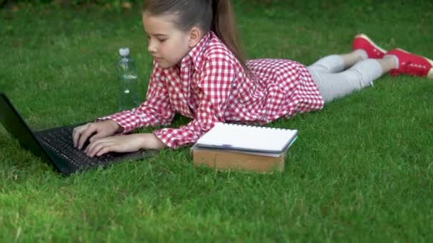 Beschikbaar 5G internet overal. Schoolmeisje in een rood shirt op groen gras in het park bij de laptop. technologie en ontwikkeling. — Stockvideo