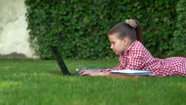 Een blank meisje, een schoolmeisje, ligt op een groen grasveld in het park en werkt op een laptop. Camera beweging, plaats voor tekst. — Stockvideo