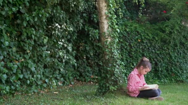 Una colegiala caucásica está sentada en el parque sobre la hierba verde. Leyendo un libro al aire libre. movimiento de la cámara. Copiar espacio. Día del libro. — Vídeos de Stock