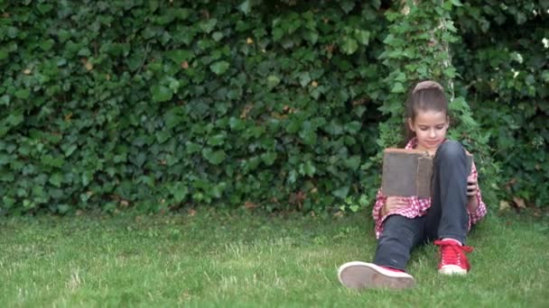 Portret uczennicy siedzi w parku na zielonym trawniku w czerwonej koszuli i czyta książkę. ruch kamery. — Wideo stockowe