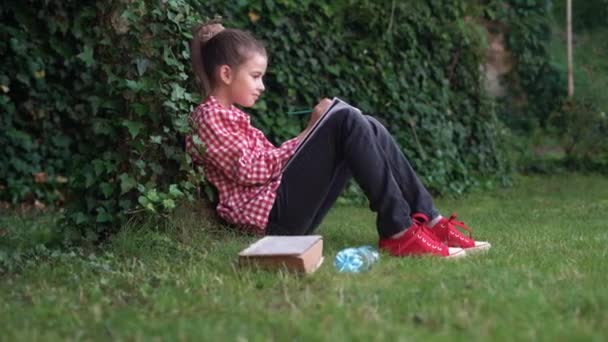 Side view of a Caucasian girl student sitting in a park near a tree holding a pencil in her hands and writing something in her notebook. Copy space. space for text. — Stock Video