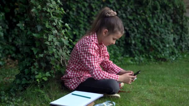 Een jong casual meisje gebruikt een mobiele telefoon in het park, een schattige tiener maakt foto 's of bekijkt een video op een moderne gadget met Wi-Fi in openbare buitenruimtes tijdens een pauze aan de universiteit. — Stockvideo