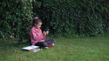 Kafesteki tişörtlü bir kız öğrenci yeşil çimenlikteki bir ağacın yanındaki parkta oturuyor. Telefonla yazı yazıyorum. Teknoloji ve kablosuz internet.