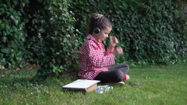 一个大学生在阳光灿烂的日子里坐在草坪上使用智能手机的画像。在大学的公园里，身穿红衫的白人女学生在手机上听着音乐. — 图库视频影像