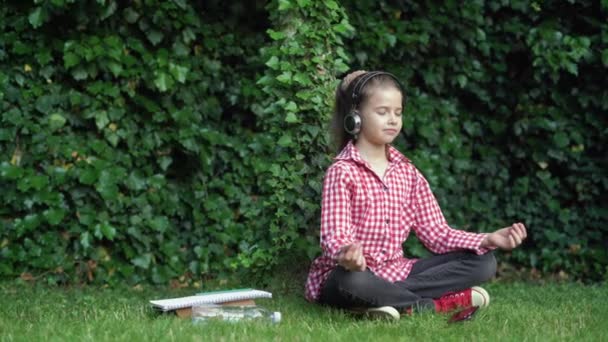 Красивая девушка отдыхает на открытом воздухе. школьница сидит на зеленой траве в позе лотоса в солнечный день. Медитация в природе. — стоковое видео