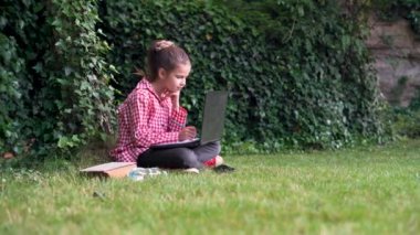 Bahar ya da yaz, kırmızı tişörtlü bir kız öğrenci yeşil çimlerde oturur, dizüstü bilgisayara bakar ve not defterindeki kalemle notlar alır. Çevrimiçi öğrenme kavramı.