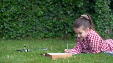 Kırmızı tişörtlü beyaz kız çimlere uzanıyor kitap okuyor ve not defterine notlar yazıyor..
