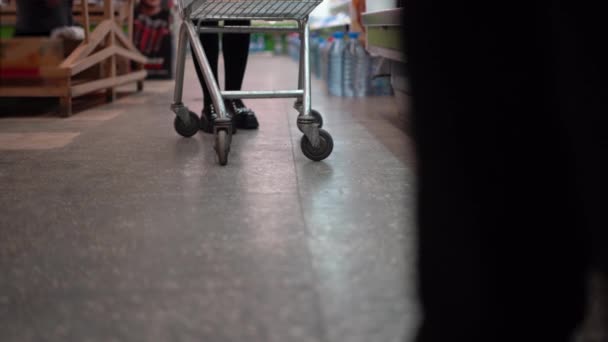 Ruedas de carro en un supermercado y piernas de mujer cerca de ellos. Sin rostro. Empuja el carro a través del hipermercado. Concepto de compra. — Vídeo de stock