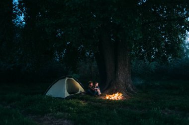 Ormanda çadırın yanındaki ateşin yanında oturan iki sevecen gezgin. Turist adam ve kız gece bir ağacın altında dinleniyorlar. Büyük bir ağaç ve alevlerin altında insanlar..