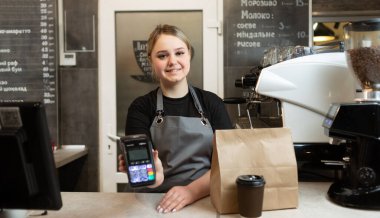 Kafeteryadaki NFS 'lerle temassız ödemeler için ödeme terminali olan Kafkasyalı barista bir kese kâğıt torba ve teslimat için bir bardak kahve hazırladı. Kafe konsepti.