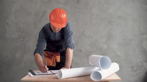 Um trabalhador da construção do sexo masculino, vestido com um uniforme e um chapéu laranja é dobrado sobre os desenhos do projeto e fazendo ajustes com um lápis. Espaço de cópia. — Vídeo de Stock