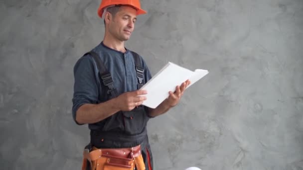 Ingenjör byggnad på en vit bakgrund. Ingenjör byggnaden håller ett papper i sina händer. En man i byggnadsarbetares uniform. Porträtt av en vuxen byggare. Byggare i grå uniform. — Stockvideo