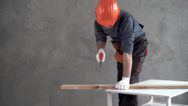 Een man gekleed in een uniform en een oranje harde hoed zaagt een blokje met een handzaag. Kopieerruimte. — Stockvideo