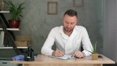 Masada oturan erkek doktor hastane ofisindeki bir belgeyle ilgili rapor yazıyor. Sağlık personeli ve hekim hizmetleri.