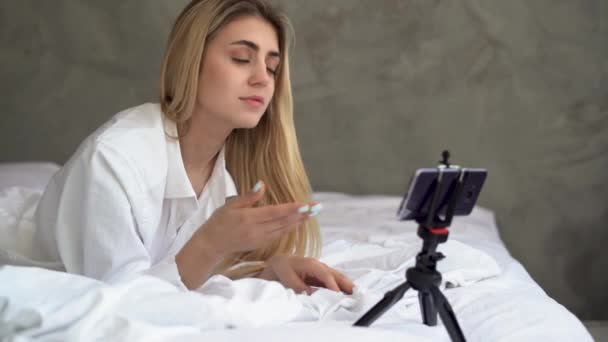 Mulher branca bonita fica em casa em sua cama faz uma chamada de vídeo através do aplicativo usando seu smartphone. Encontra notícias interessantes, emoções de surpresa. — Vídeo de Stock