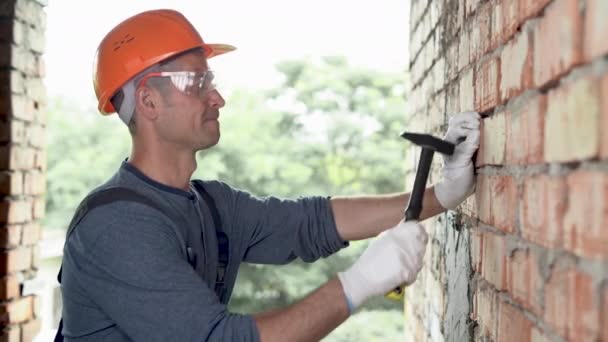 Un hombre constructor trabaja en un sitio de construcción, martillando un clavo de metal en una pared de ladrillo rojo con un martillo. Vestido con uniforme, gafas y casco de construcción naranja. — Vídeos de Stock