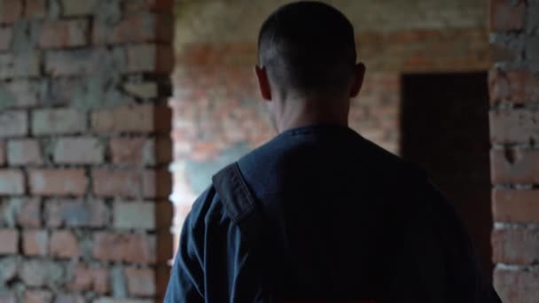 Vista trasera de un hombre constructor vestido con un uniforme camina a lo largo de un sitio de construcción de una pared de ladrillo. — Vídeos de Stock