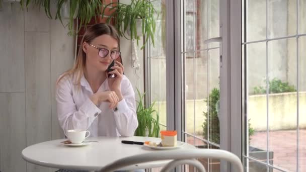 창문 근처 카페의 테이블에는 안경을 쓴 젊은 금발의 여성 이 스마트폰으로 대화를 나누고 있다. 옆에 커피 한 잔이요. 복사 공간. — 비디오