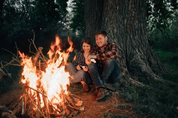 Close-up lovers are sitting by the fire and frying marshmallows.Camping in the forest.Enamored guy and girl travelers are preparing food.Outdoor tourism and recreation concept.A tree and people under