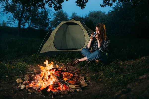 girl traveler drinks tea sits next to a tent in the forest in the evening. Camping woman resting in nature drinks coffee from a metal mug. Travel and drinks concept.