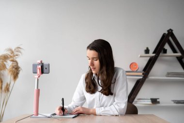 Masada oturan konsantre kız öğrenci bir not defterine üç ayaklı bir telefonla notlar alır. Kız ev ödevini cep telefonundan yazar. Çevrimiçi eğitim ve aygıt. Metin için yer