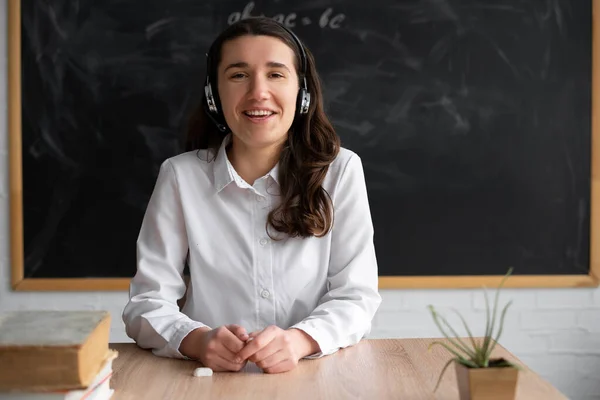 portrait of a happy teacher or tutor sitting in the classroom at the table teaches a lesson online in headphones, headset. Chalk board in the background. Lecture on the Internet. Place for text.