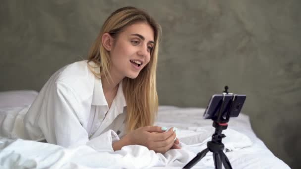 Élégant jeune femme se trouve le matin sur le lit dans une chemise mans fait un appel vidéo à travers son smartphone en utilisant l'application émotion de plaisir sur son visage. — Video