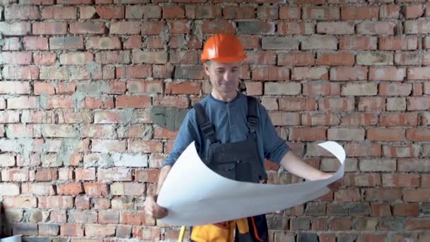 Un hombre constructor, vestido con un sombrero duro y uniforme naranja, y gafas, se levanta contra una pared de ladrillo y mira una gran hoja de papel. El concepto es estudiar el plan de construcción. — Vídeos de Stock