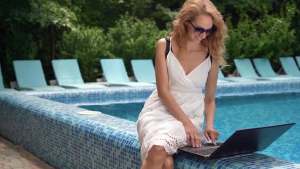 Uma mulher com um portátil na piscina. Trabalho remoto e conceito nômade global. Freelancer jovem no resort. Teleconferência e trabalho freelance. — Vídeo de Stock
