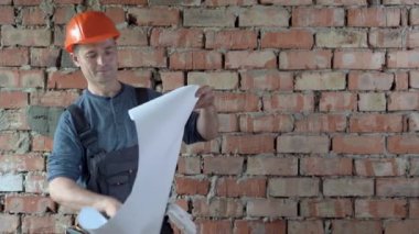Beyaz bir erkek müteahhit, turuncu bir şapka takmış kırmızı tuğla bir duvarın arka planında duruyor ve üniforma binanın çizimlerini içeren bir çarşaf seriyor. yenileme konsepti.