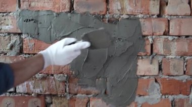 Eldivenli usta bir müteahhidin ellerine yakın çekim yapar, bir spatula tutar ve tuğla duvara sıva sürer..