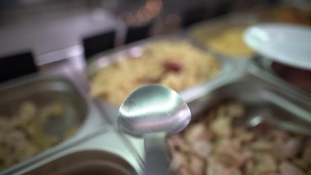 Close-up van een grote lepel pikt spiraalvormige pasta met gerookte worsten op uit een grote container op de foodserveerlijn in een buffet en giet in een bord. Van de eerste persoon. — Stockvideo