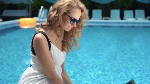 Young beautiful caucasian woman is sitting near the pool and working at her laptop. — Stock Video