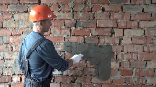Biały mężczyzna budowniczy stoi w pobliżu cegły ściany i nakłada cement na ścianę łopatką. Ubrany w mundur roboczy i ochronny, pomarańczowy, twardy kapelusz. — Wideo stockowe