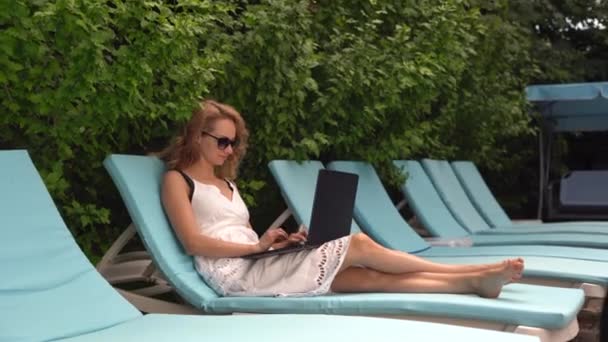 Uma mulher com um portátil junto à piscina. Sentado em uma espreguiçadeira e usando um laptop na piscina durante as férias. O conceito de trabalho remoto em qualquer lugar do mundo. — Vídeo de Stock