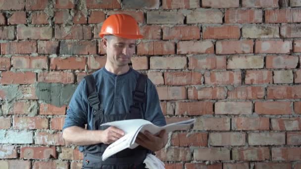 Um construtor masculino caucasiano vestido com um capacete laranja e um uniforme está contra uma parede de tijolos, segura plantas com um projeto de casa em suas mãos e as estuda cuidadosamente. espaço de cópia. — Vídeo de Stock