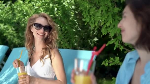 Podczas wakacji, dwie piękne, kaukaskie damy w okularach przeciwsłonecznych siedzą na leżakach w pobliżu basenu, pijąc koktajl ze szklanek i rozmawiając słodko. — Wideo stockowe