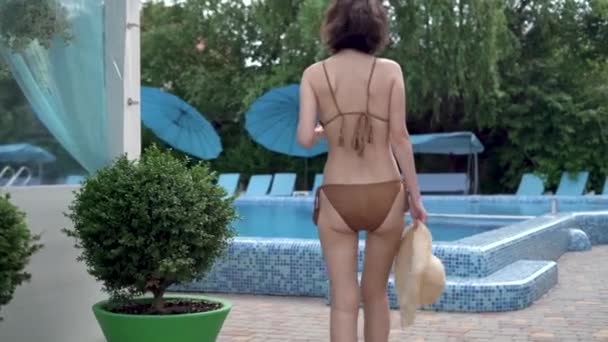 Вид ззаду одна красива молода жінка, одягнена в купальник, тримає солом'яний капелюх в руках, йде біля великого басейну. Концепція відпустки на курорті, відпочиваючи в готелі з — стокове відео