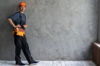 Turuncu bir şapka ve alet kemeri takan bir inşaatçı gri bir duvarın yanında duruyor. Beton duvar ve inşaatçı. Mekanın yakınındaki tamircide mesaj var. İnşaat alanı konsepti. İç duvar yenilemesi.