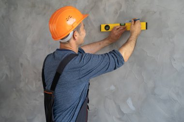 Miğferli bir ustabaşının duvar ölçüsünü bir seviyeyle ölçtüğü dikiz görüntüsü. Bir inşaat işçisi duvarın durumunu kontrol eder. Kontrolör, efendinin işini devralır. - Ev. Metin için yer
