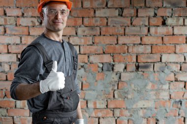 Bir inşaat mühendisi ya da mimar koruyucu kasklı ve gözlüklü içeride tuğla bir duvara karşı duruyor ve parmağını kaldırıyor. Erkek ustabaşı. Metin için boşluk. Yenileme konsepti