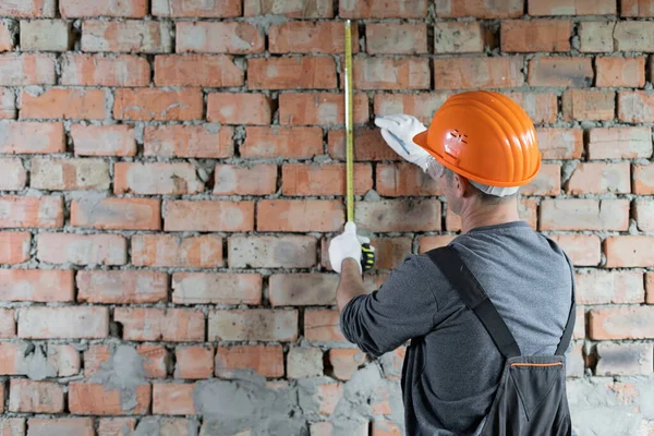 builder in a protective helmet and goggles measures the height on the wall with a metal tape measure and marks the level with a pencil. Brick wall and architect . Renovation concept. Place for text