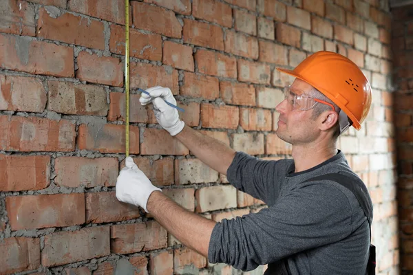 profile view of a civil engineer in a protective helmet and glasses measures a brick wall with a tape measure at a construction site. Measurements of the height of the wall. Construction site concept